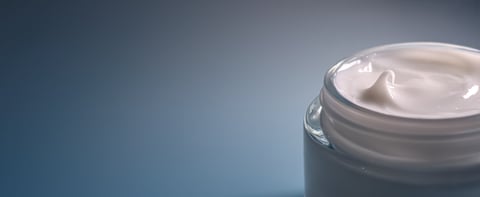 White cosmetic cream in an open glass jar, showing a smooth texture against a subtle blue background, focusing on hydration and self care